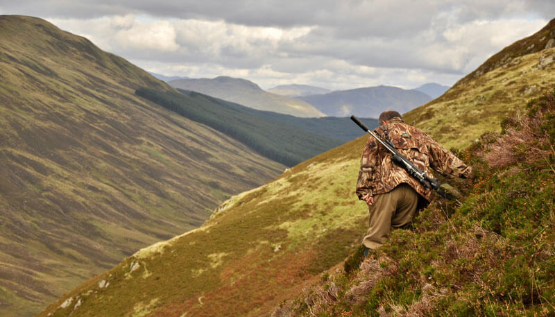 A man walking through the hills on a hunting trip looking for deer and carrying a rifle.